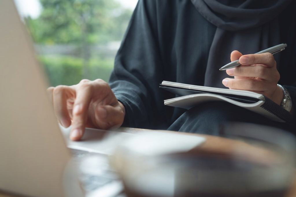 Muslim woman Person writing ideas in a notebook while hesitating to take action on a laptop The Firdaws Connection